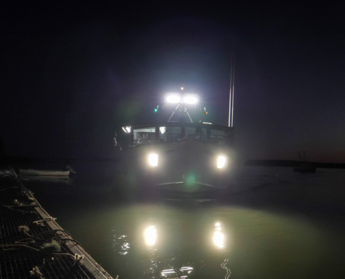 Front view of a boat by a dock at night, lights on flashing at camera